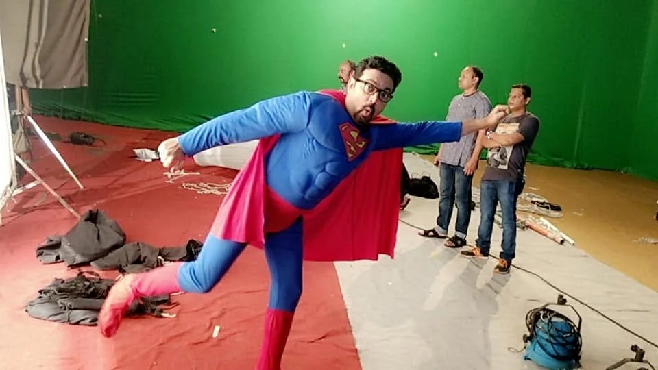 Abhinandan in a blue superhero costume and red cape, between takes on a green-screen set.
